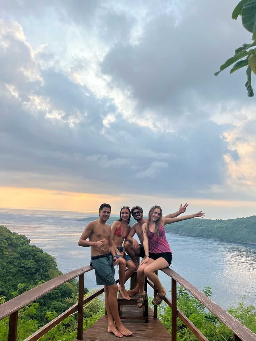 Four people posing on a wooden viewpoint overlooking Crystal Bay, Nusa Penida, at sunset with lush green cliffs and calm ocean in the background