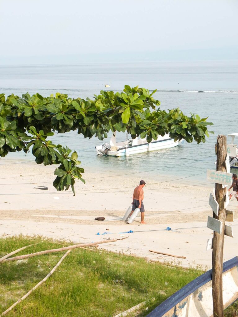 A diver carrying a scuba tank along the beach at Toya Pakeh, Nusa Penida, with a small boat moored in the calm shallow water
