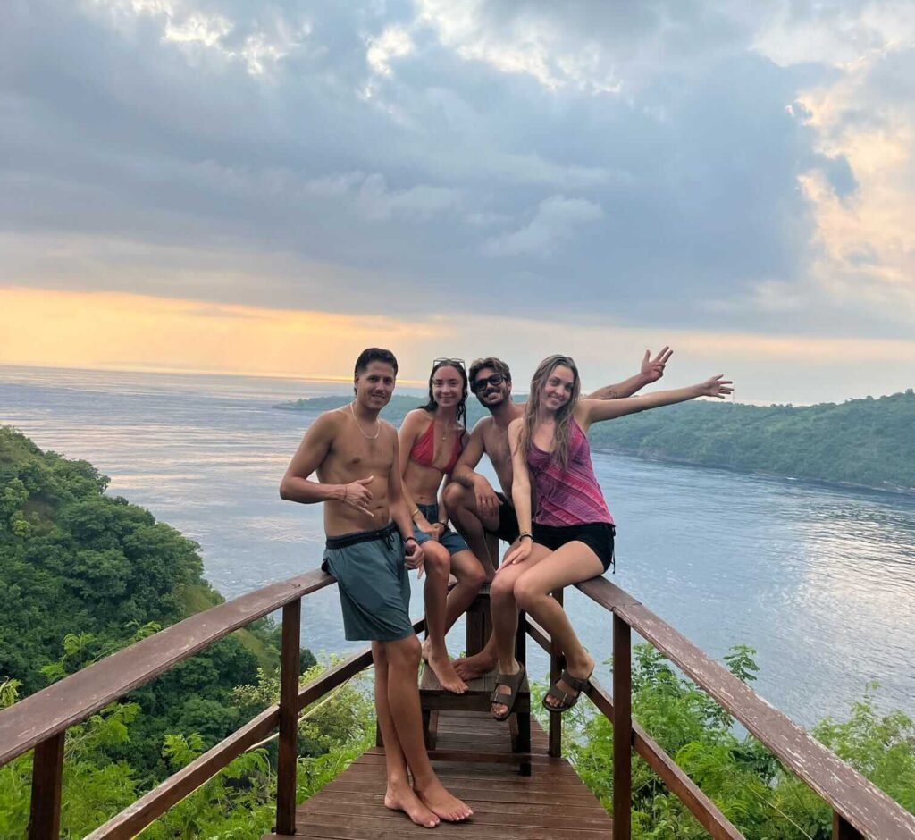 Four people posing on a wooden viewpoint overlooking Crystal Bay, Nusa Penida, at sunset with lush green cliffs and calm ocean in the background